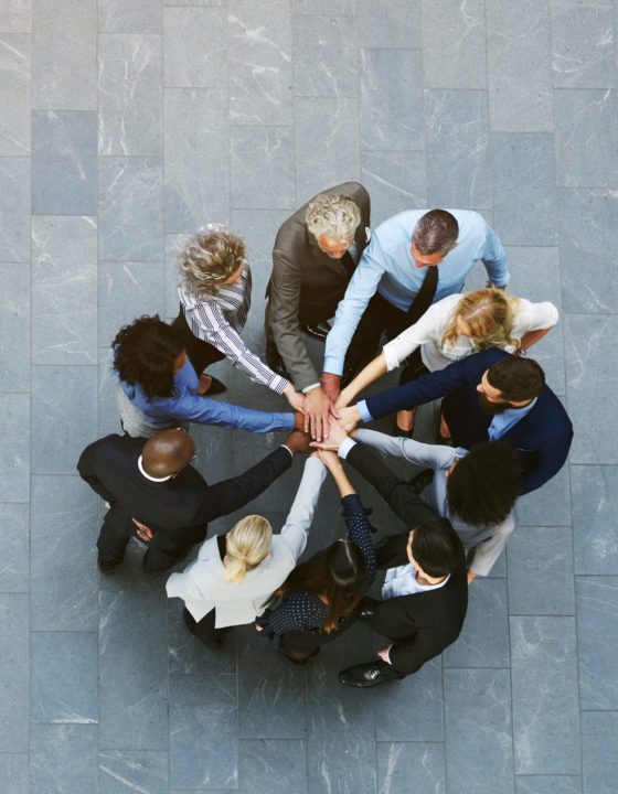 From above black and white office working people having teambuilding in hall standing with hands stacked.